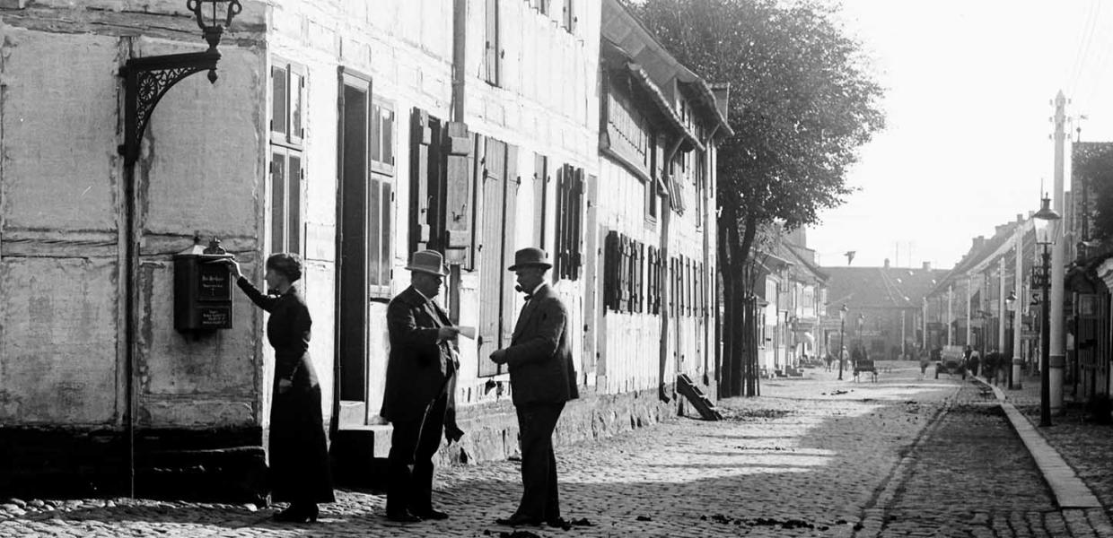 Eine Frau steht daneben, und zwei Männer mit Melone unterhalten sich im Trægården – in alten Zeiten