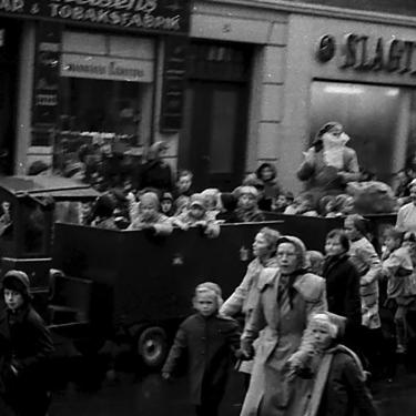 Ein großes Auto mit Kobolden fährt an den Geschäften in der Hauptstraße vorbei - im Jahr 1955